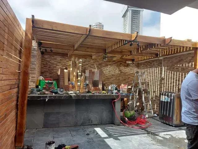Patio exterior con paredes de ladrillo, pérgola de madera, mostrador y herramientas, con una persona sentada a la derecha.