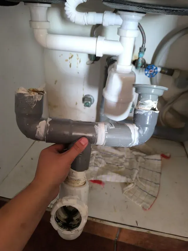 Una mano sostiene un tubo de PVC gris debajo de un fregadero, con los accesorios de desagüe a la vista y las tuberías blancas.