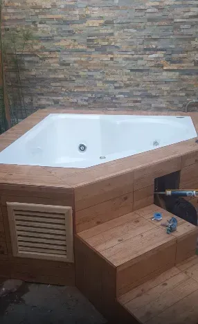 Bañera de hidromasaje blanca en esquina con revestimiento de madera y escalones en un baño alicatado.