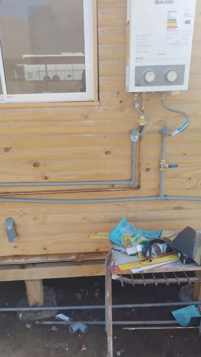 Pared revestida de madera con contadores de servicios públicos, tuberías a la vista y una pequeña mesa desordenada debajo.