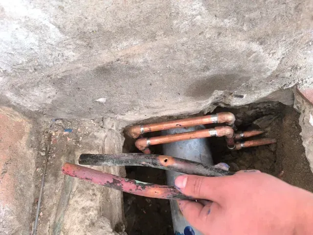 Una mano sostiene una herramienta de metal oxidada junto a tuberías de cobre dentro de una abertura de hormigón rugoso.