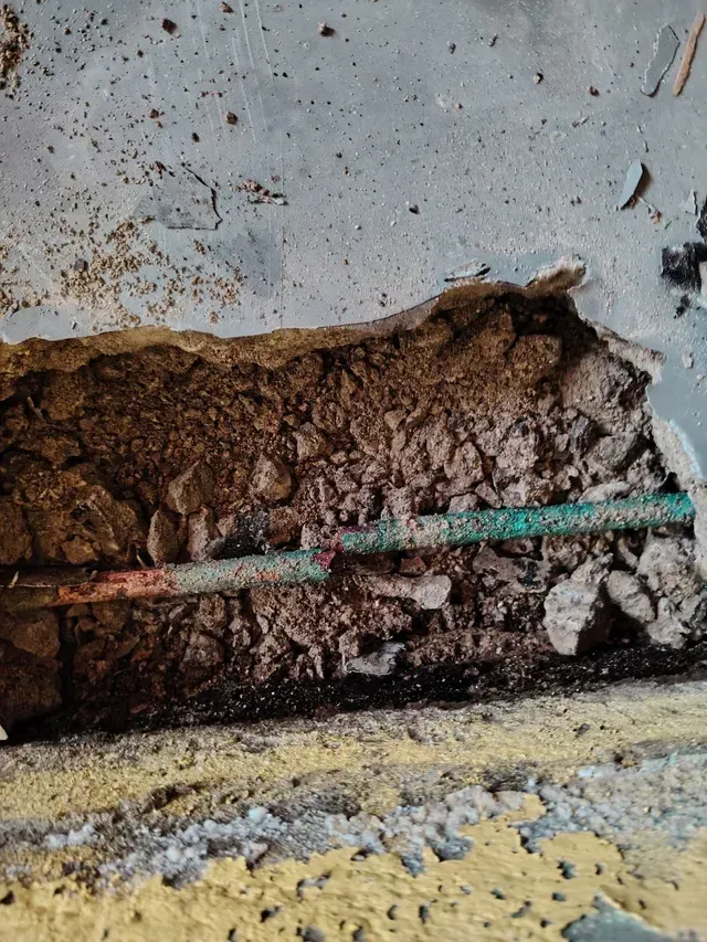 Primer plano de una pared dañada que muestra yeso marrón expuesto, una tubería verde y escombros desmoronándose a lo largo de la base.