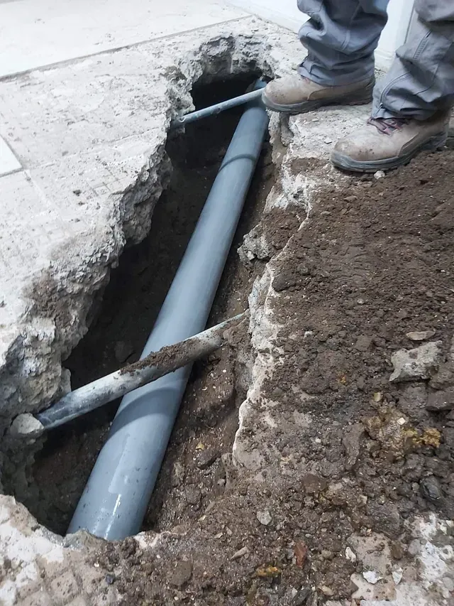 Zanja abierta con una tubería gris expuesta entre hormigón y tierra, con botas de trabajadores cerca.