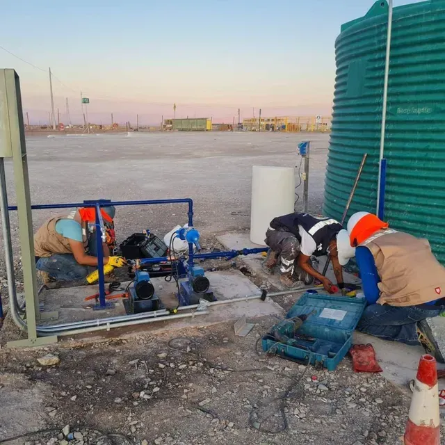 Obreros reparando tuberías junto a un tanque verde en una polvorienta obra en construcción al amanecer.