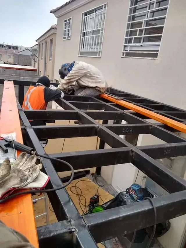 Dos trabajadores instalan una estructura de acero negro en una azotea junto a la pared de un edificio.