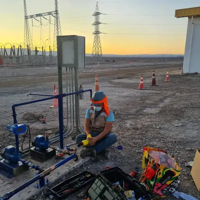 Trabajador con chaleco naranja y casco trabajando en tuberías azules en una planta de servicios públicos cerca de líneas eléctricas al atardecer.