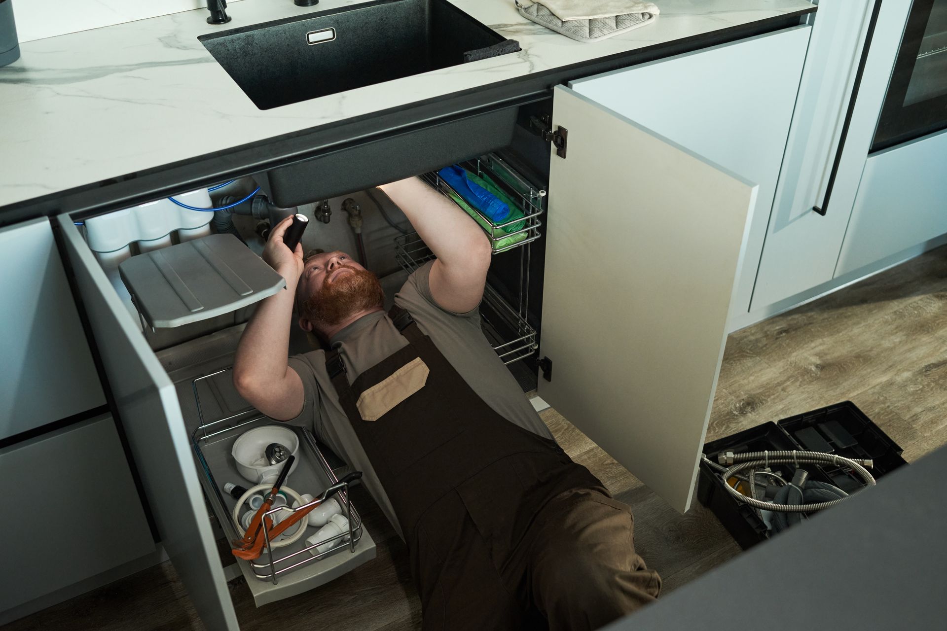 A plumber lying on the floor under the kitchen sink repairing pipes with tools and equipment nearby