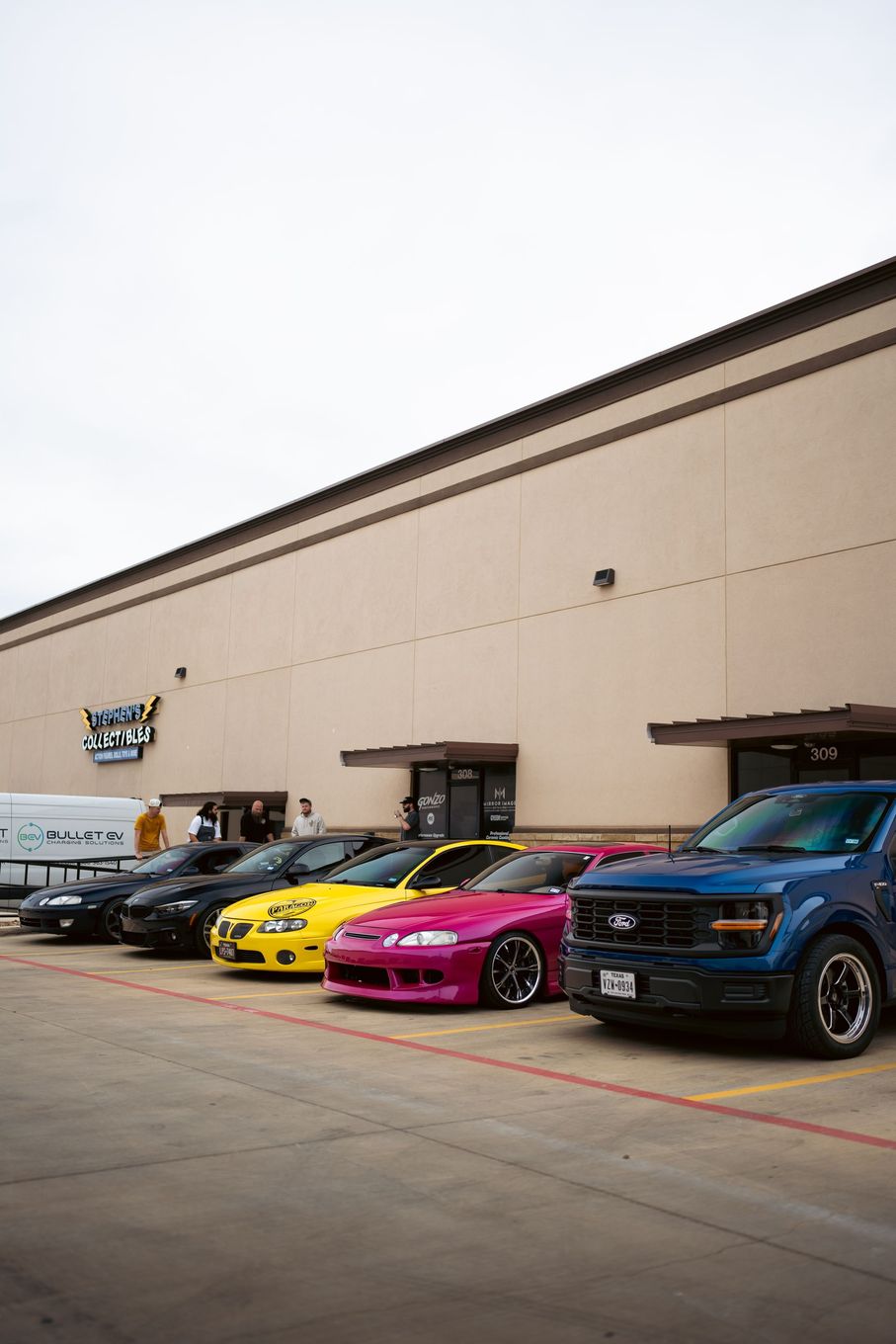 Four cars parked outside a beige warehouse, including a yellow, pink, and blue vehicle.