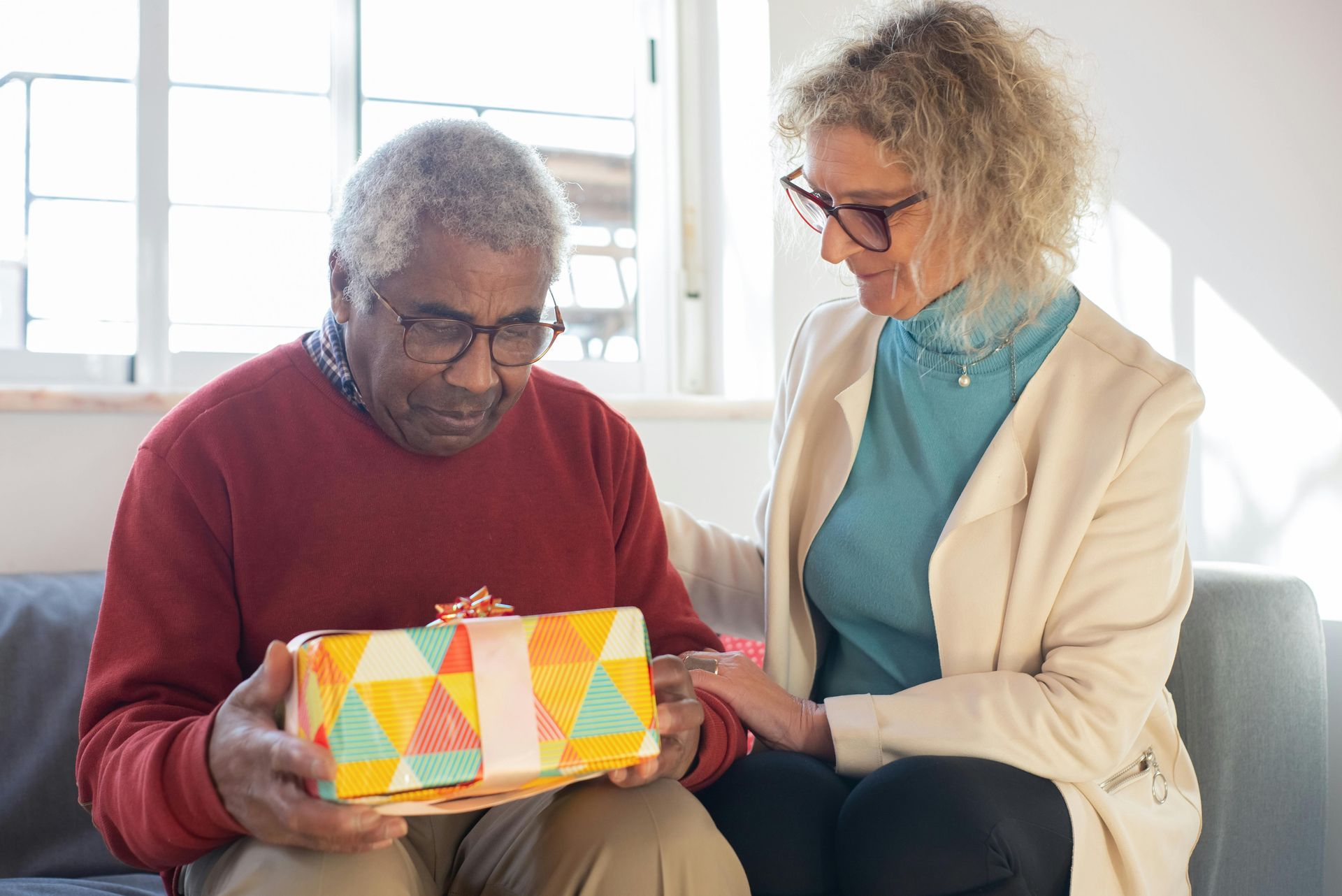 A woman is giving an older man a gift while sitting on a couch.