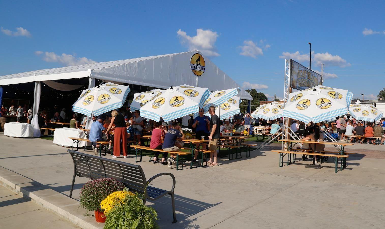 Outdoor beer garden with tables, umbrellas, and people under a large tent on a sunny day.