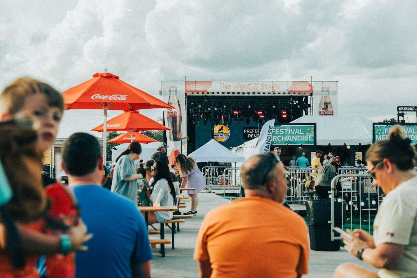 Festival crowd watching a band on stage, orange umbrellas and picnic tables in view.