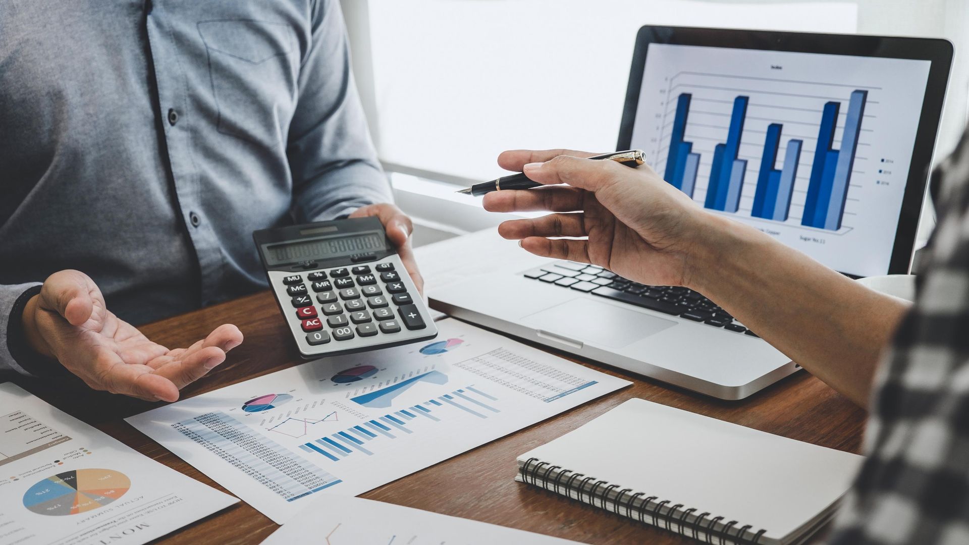 Two people reviewing financial data with a calculator, laptop, and charts on a desk.