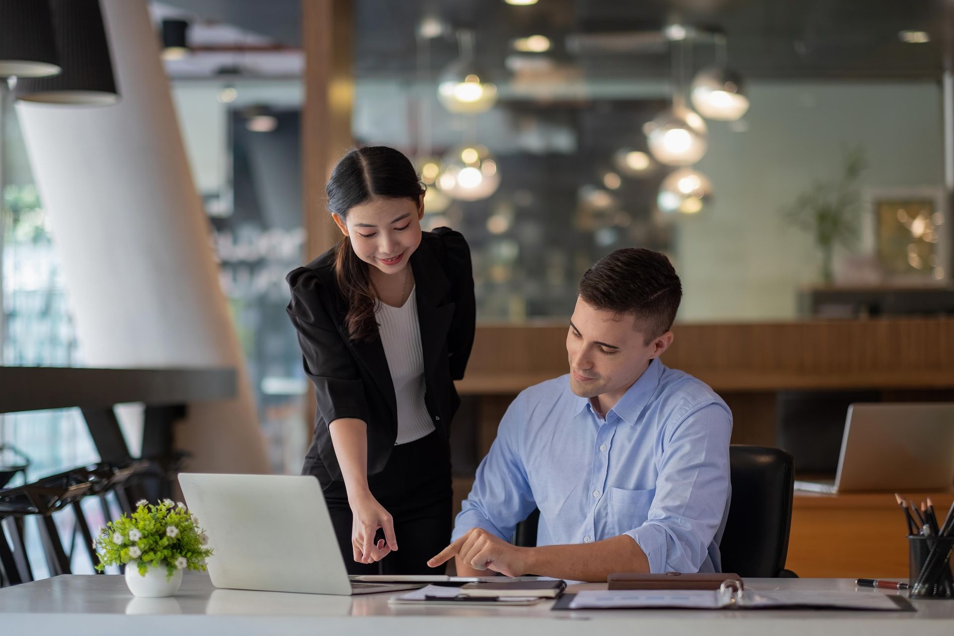 Woman pointing at a laptop, assisting a man seated at a desk in an office.