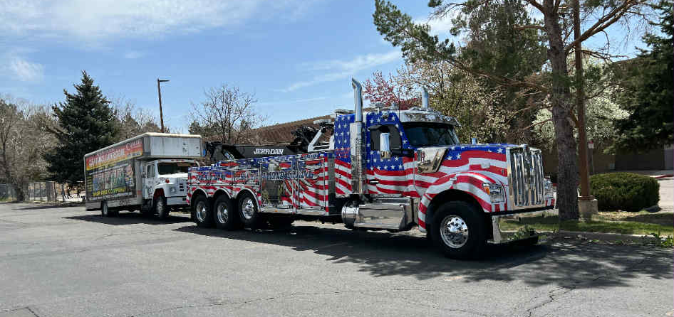 Patriotic tow truck pulling a box truck, parked on a paved lot on a sunny day.
