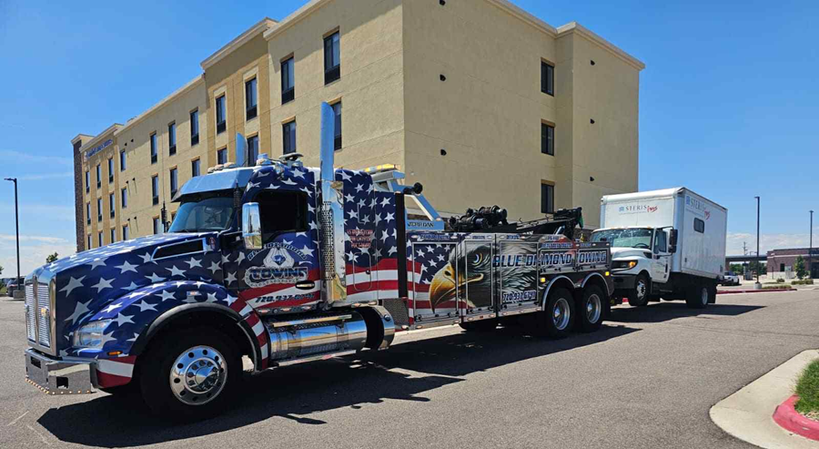 Patriotic tow truck pulling a white box truck on a sunny day in front of a hotel.