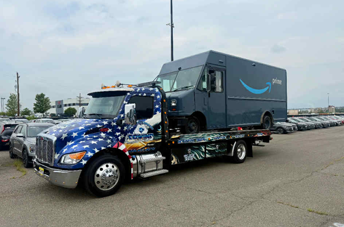 Tow truck with American flag design carrying a gray Amazon delivery van in a parking lot.