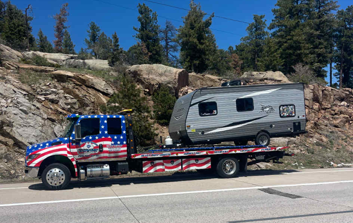Tow truck with American flag paint hauling a travel trailer on a highway near rocky terrain.