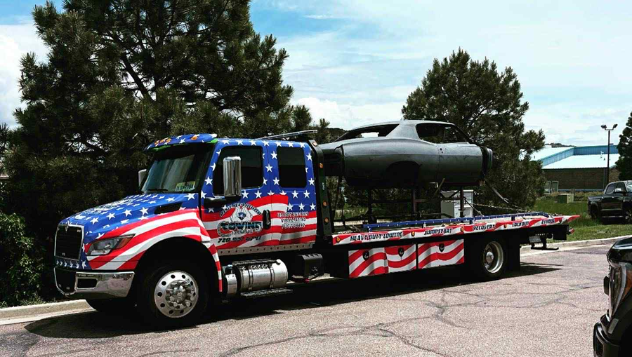 Tow truck with an American flag design hauling a dark-colored airplane fuselage.