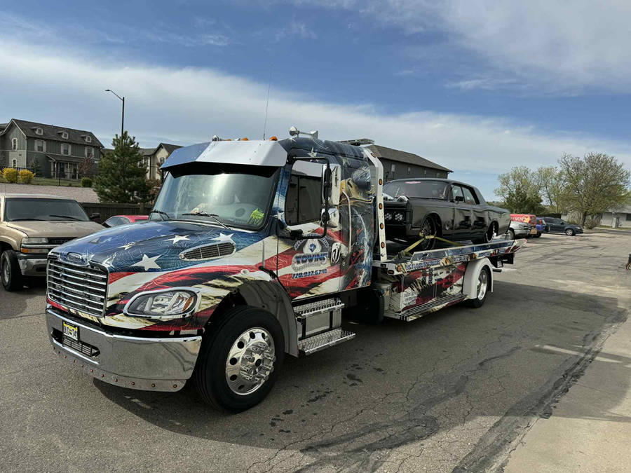 Tow truck with American flag design carrying a classic car on a sunny day.