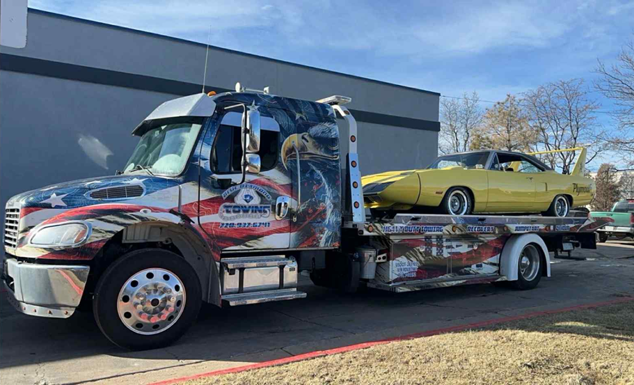 Yellow classic car on a flatbed tow truck with American flag graphics against a blue sky.