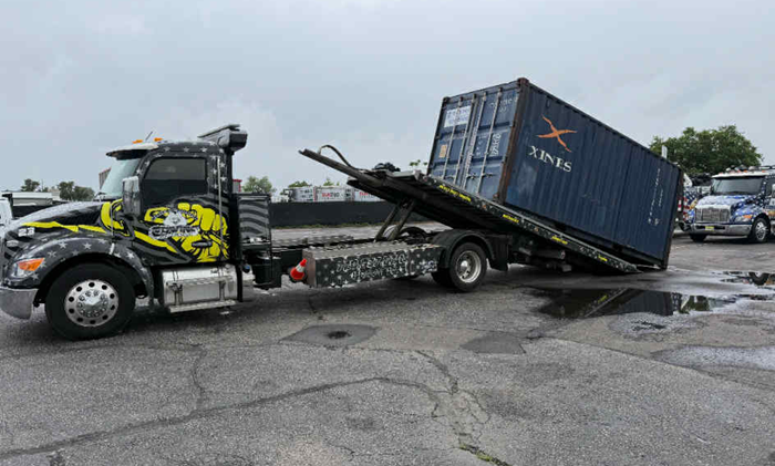 Tow truck lifting a blue shipping container on a gray paved area. The truck has black and yellow design.