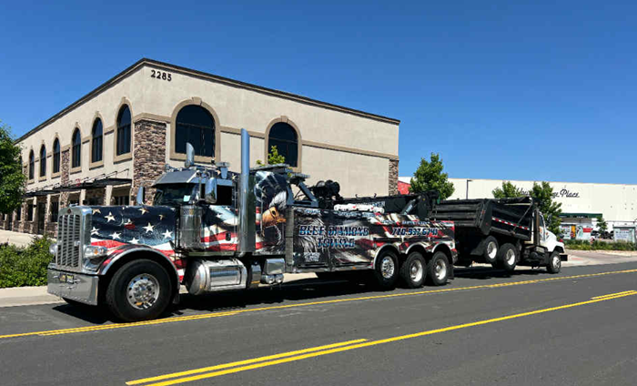 American flag-themed tow truck towing a dump truck on a sunny street, in front of a building.
