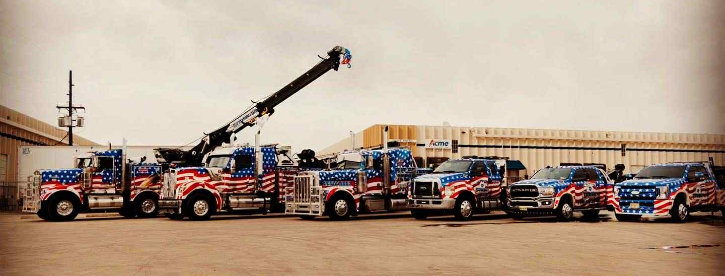 A row of tow trucks and service vehicles with an American flag theme parked outside on a cloudy day.