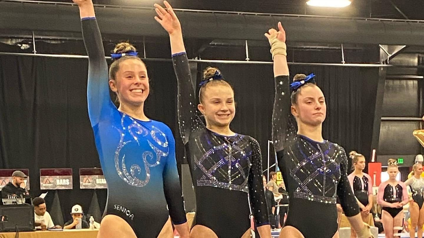 Three female gymnasts are standing next to each other with their arms in the air.
