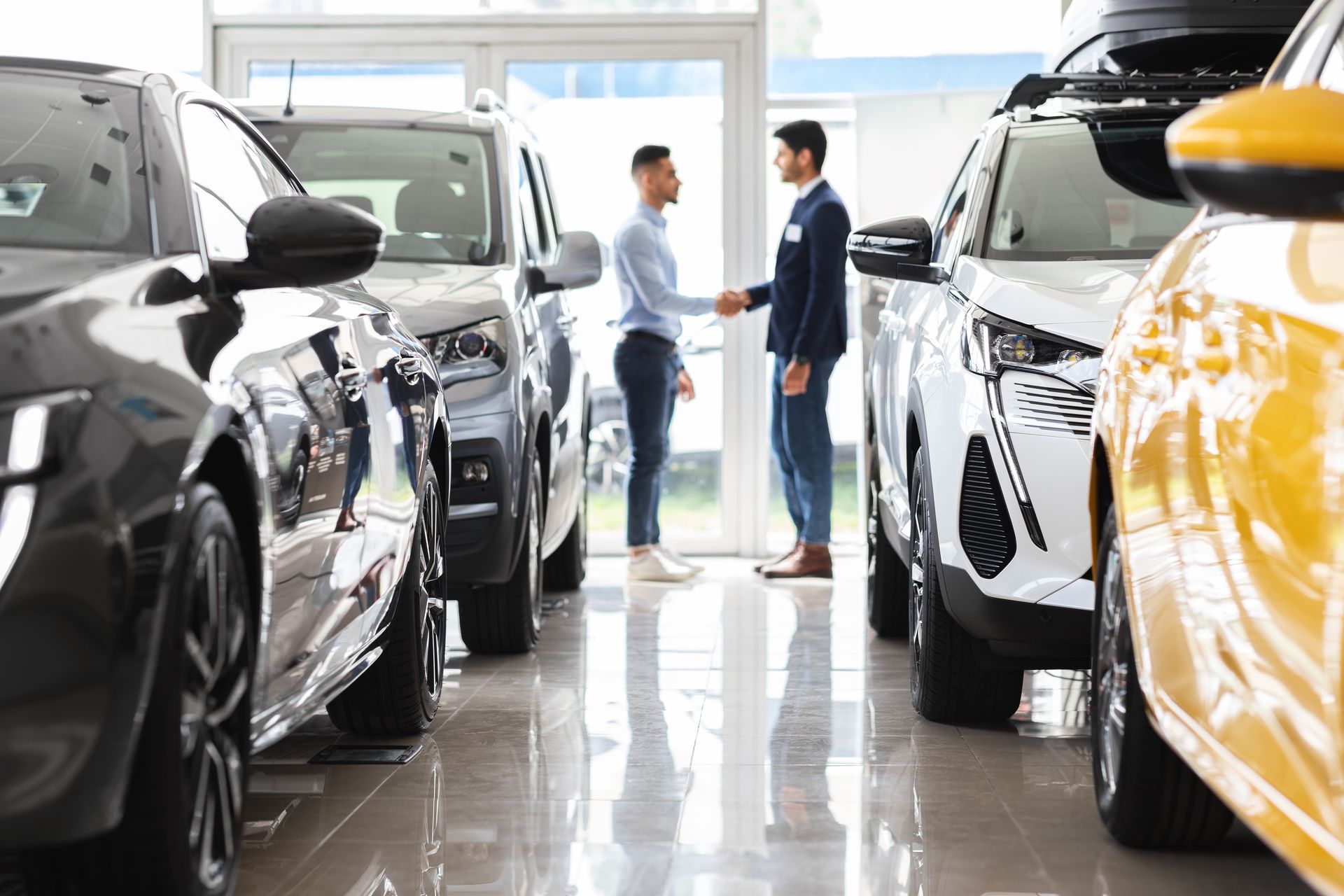 Two men shaking hands in a car dealership, surrounded by new vehicles.