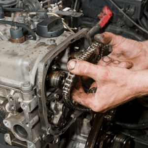 Manos reparando el motor de un coche, sujetando una cadena de distribución dentro del compartimento del motor abierto.