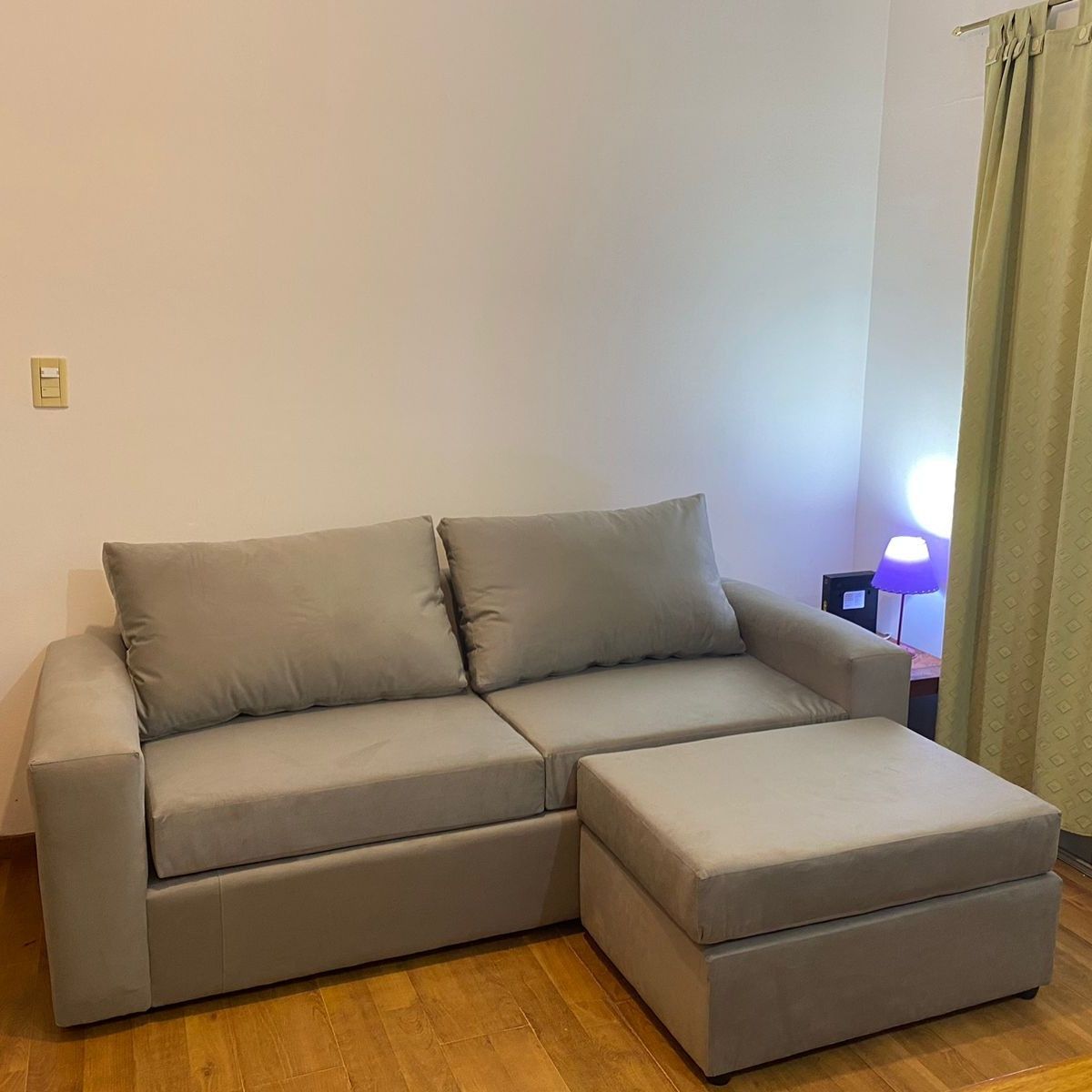 Sofá y otomana beige en una habitación con paredes de color claro y suelo de madera, junto a una cortina.