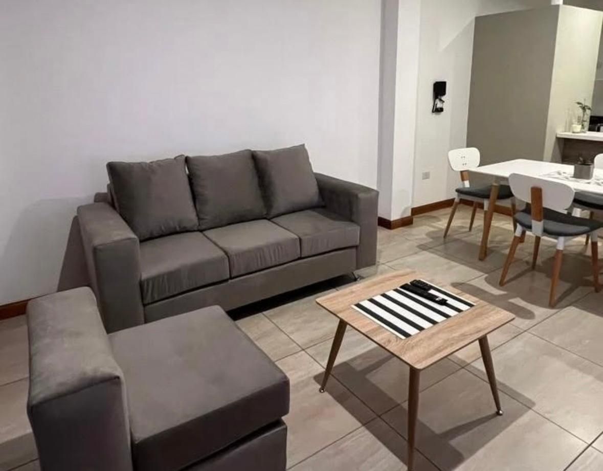 Sala de estar con sofá gris, silla, mesa de centro cuadrada y mesa de comedor.