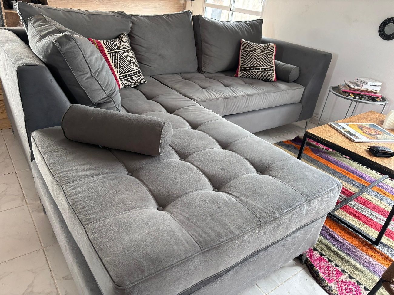 Sofá seccional gris con cojines capitoné, almohadas y una pequeña mesa auxiliar en una sala de estar.