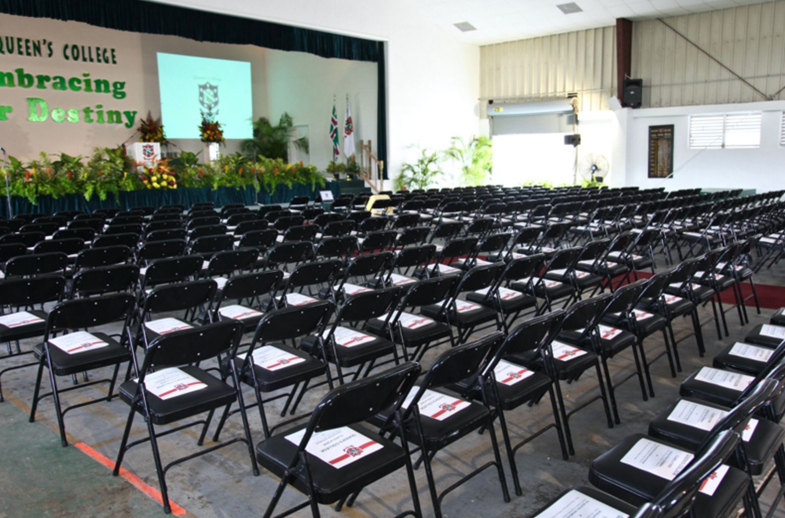 Rows of black folding chairs are lined up in front of a sign that says queen 's college embracing destiny