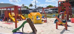 A playground with a slide , swings , and a house in the sand.