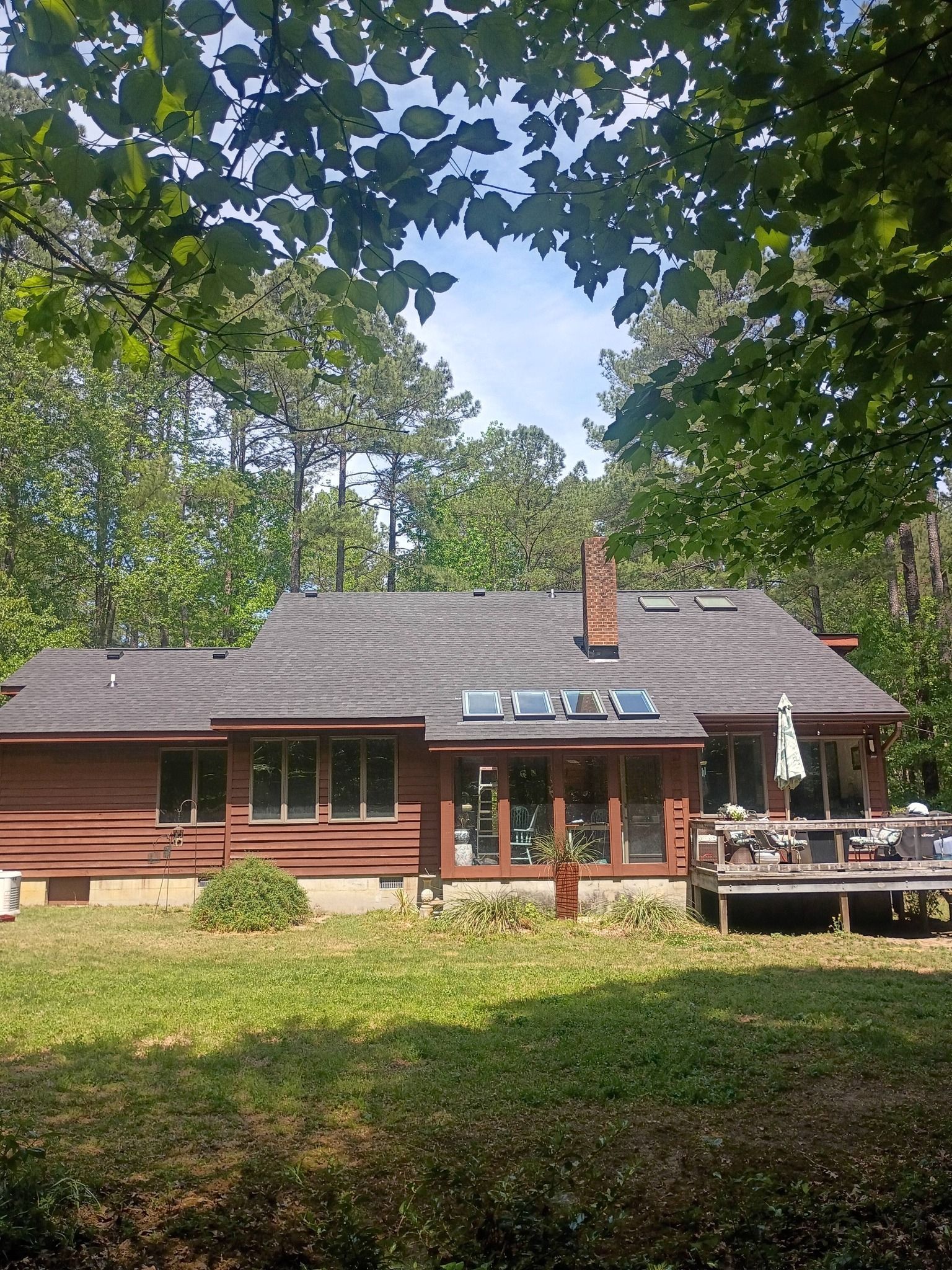 Red wooden cabin with a porch and chimney, partly shaded by trees in a green wooded yard