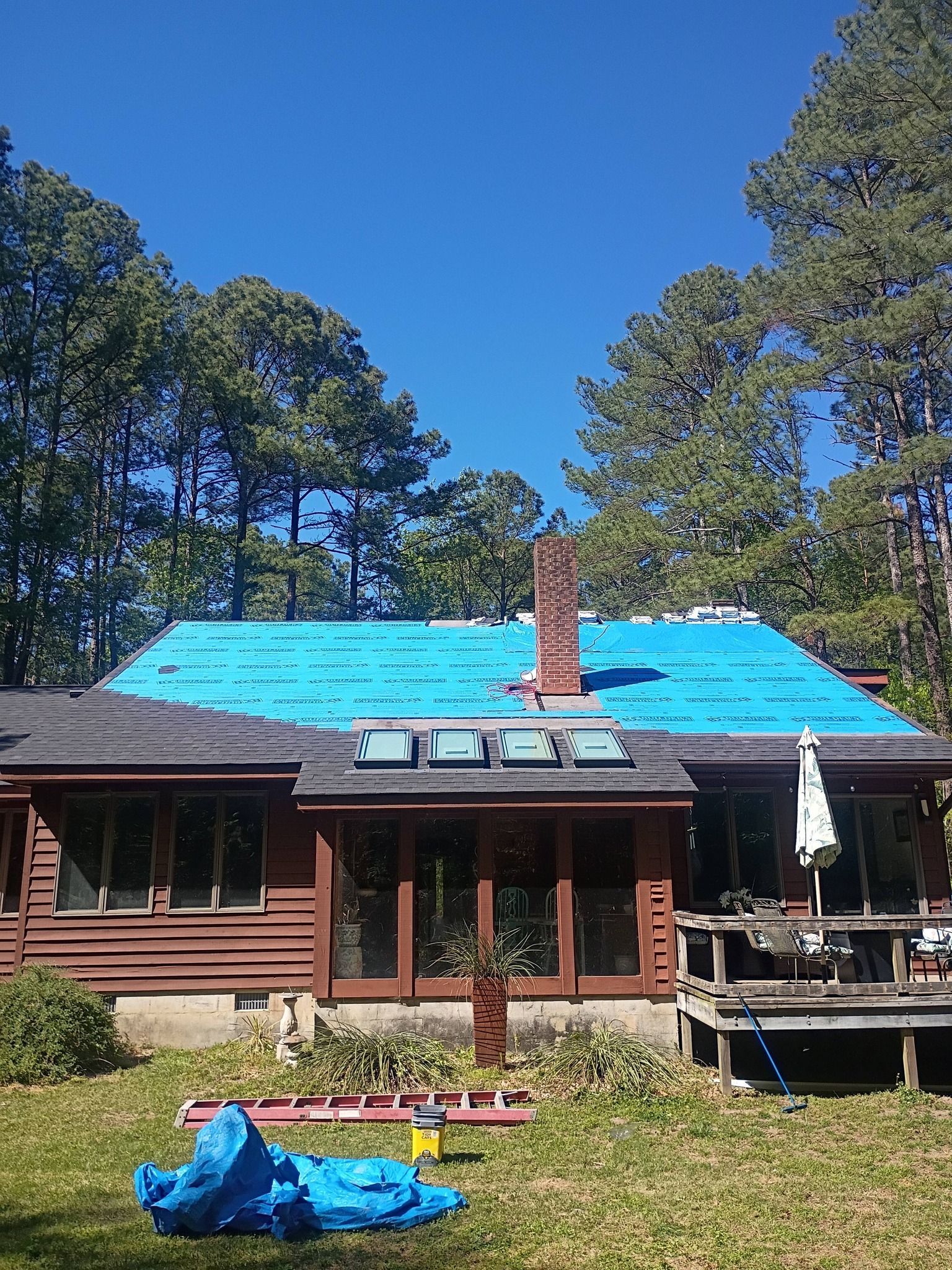 Wooden house with bright blue roof tarp, brick chimney, and deck in a sunny wooded yard