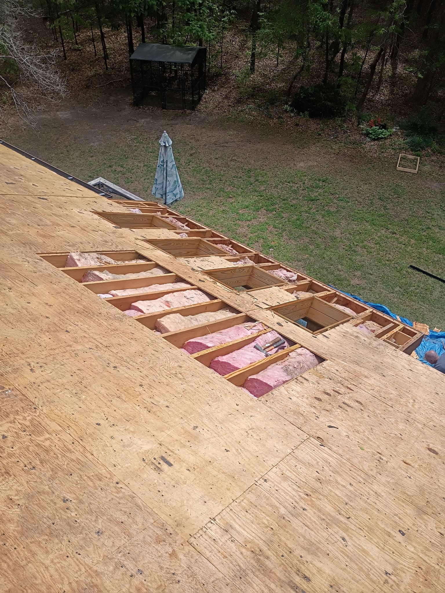 Roof under construction with exposed plywood, roofing materials, and a small blue tarp near a grassy yard.