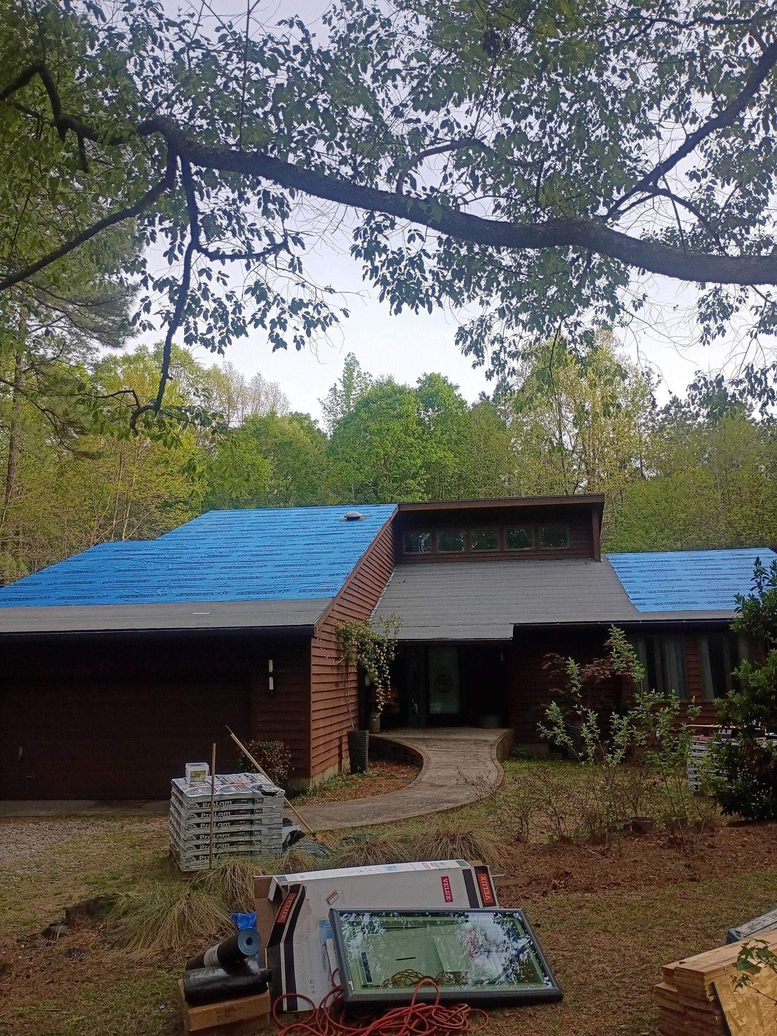 House with blue tarp-covered roof and front porch, seen through trees; car parked in foreground.