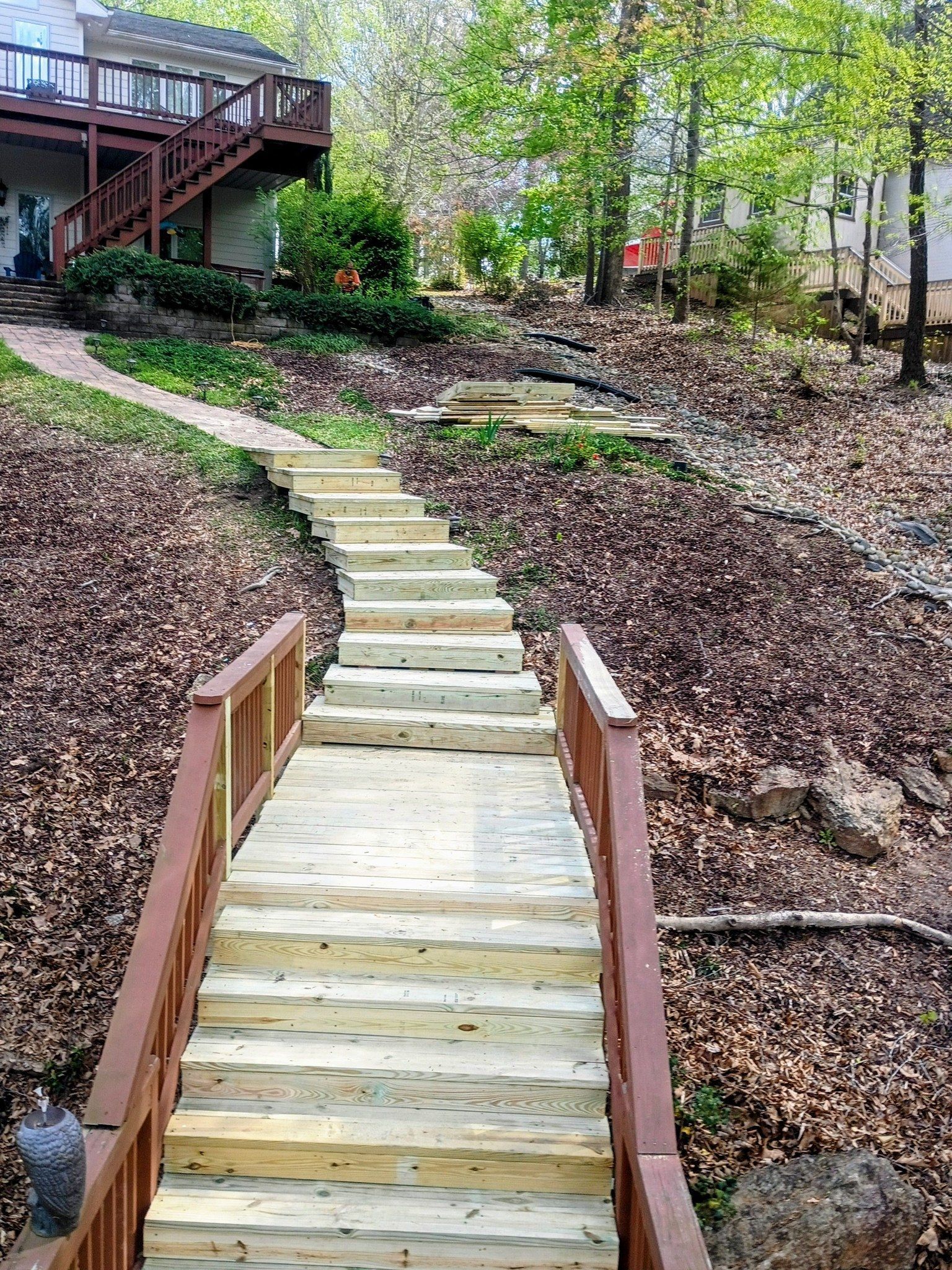Wooden staircase up a leafy hillside to a deck, with railings and a house on the left.