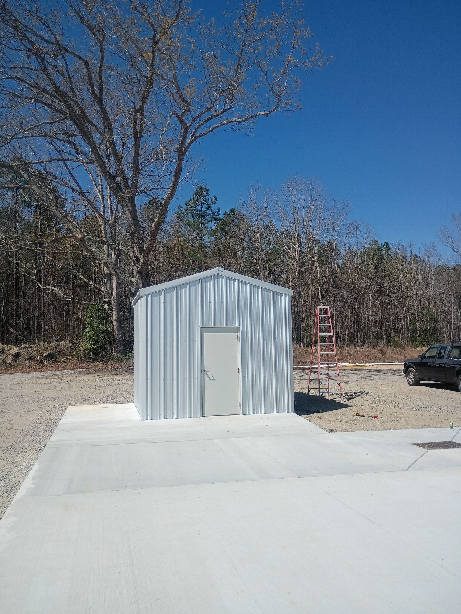Small gray metal shed beside a concrete pad, with trees and a blue sky in the background.