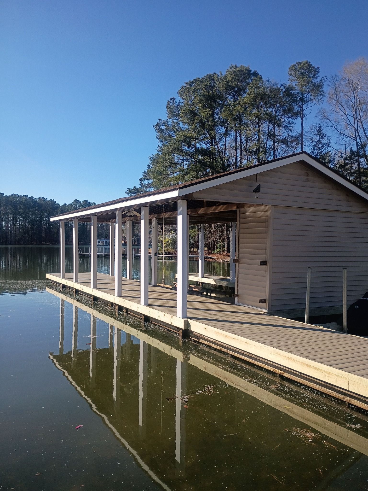 Covered dock on a calm lake, with white posts reflecting in the water under a clear blue sky.