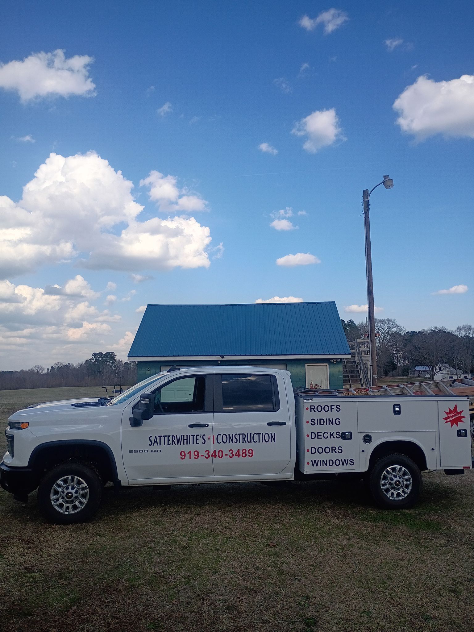 White service truck parked in a field under a blue sky, with a small blue-roofed structure behind it.