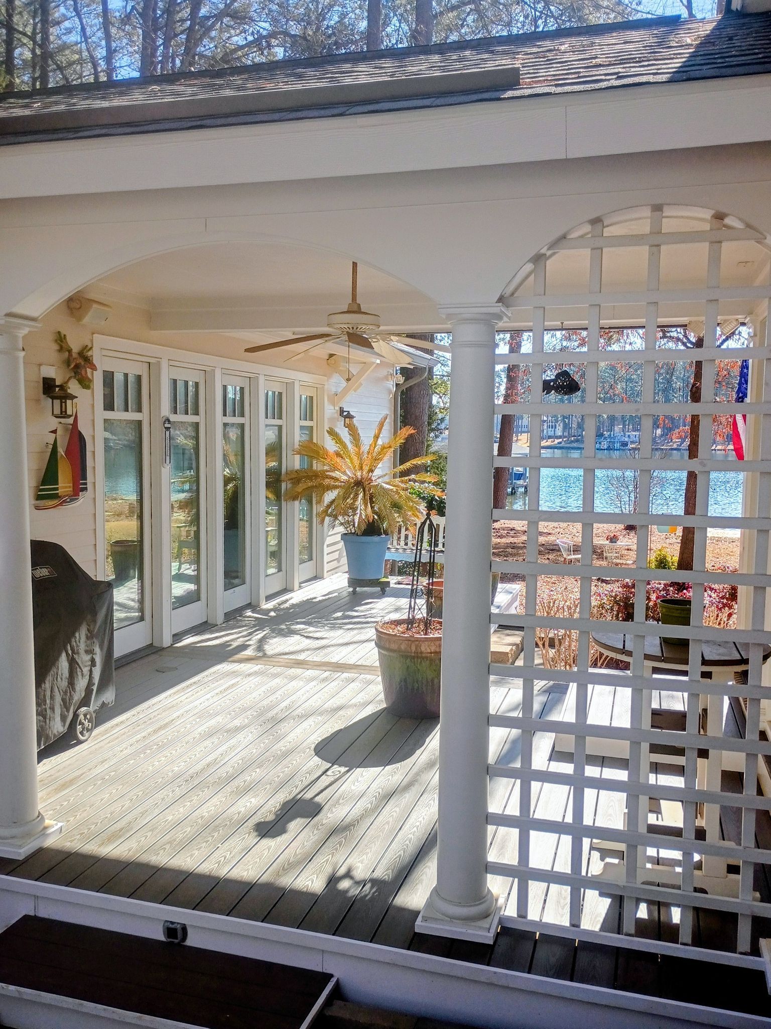 Sunlit screened porch with white columns, lattice wall, glass doors, and potted plants.