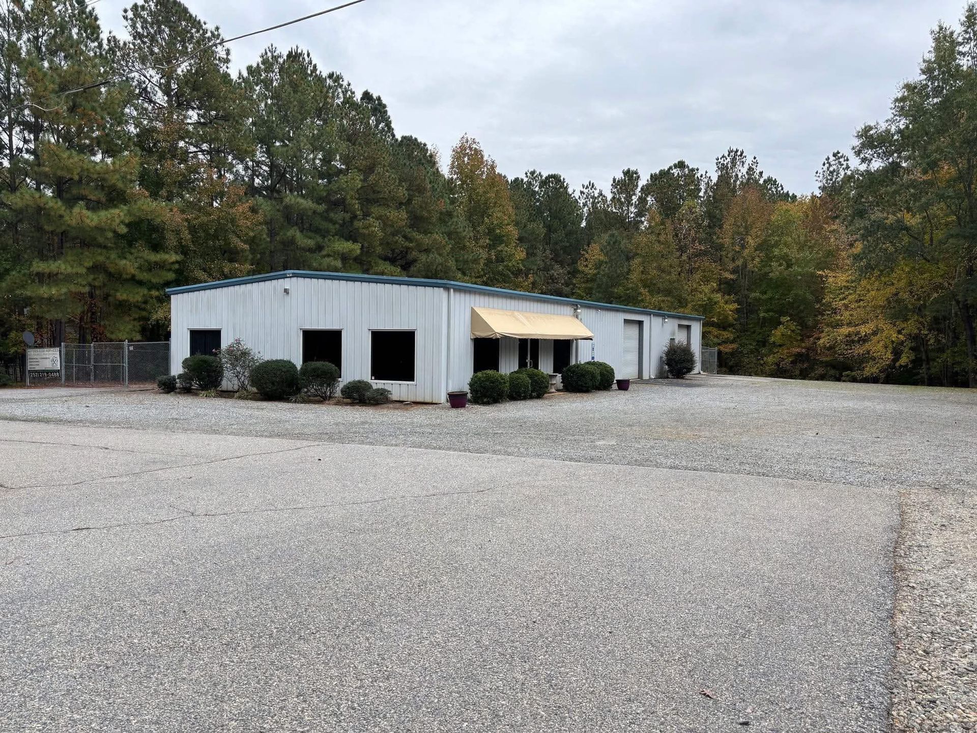 White industrial building with green awning on a gravel lot, surrounded by trees under cloudy sky
