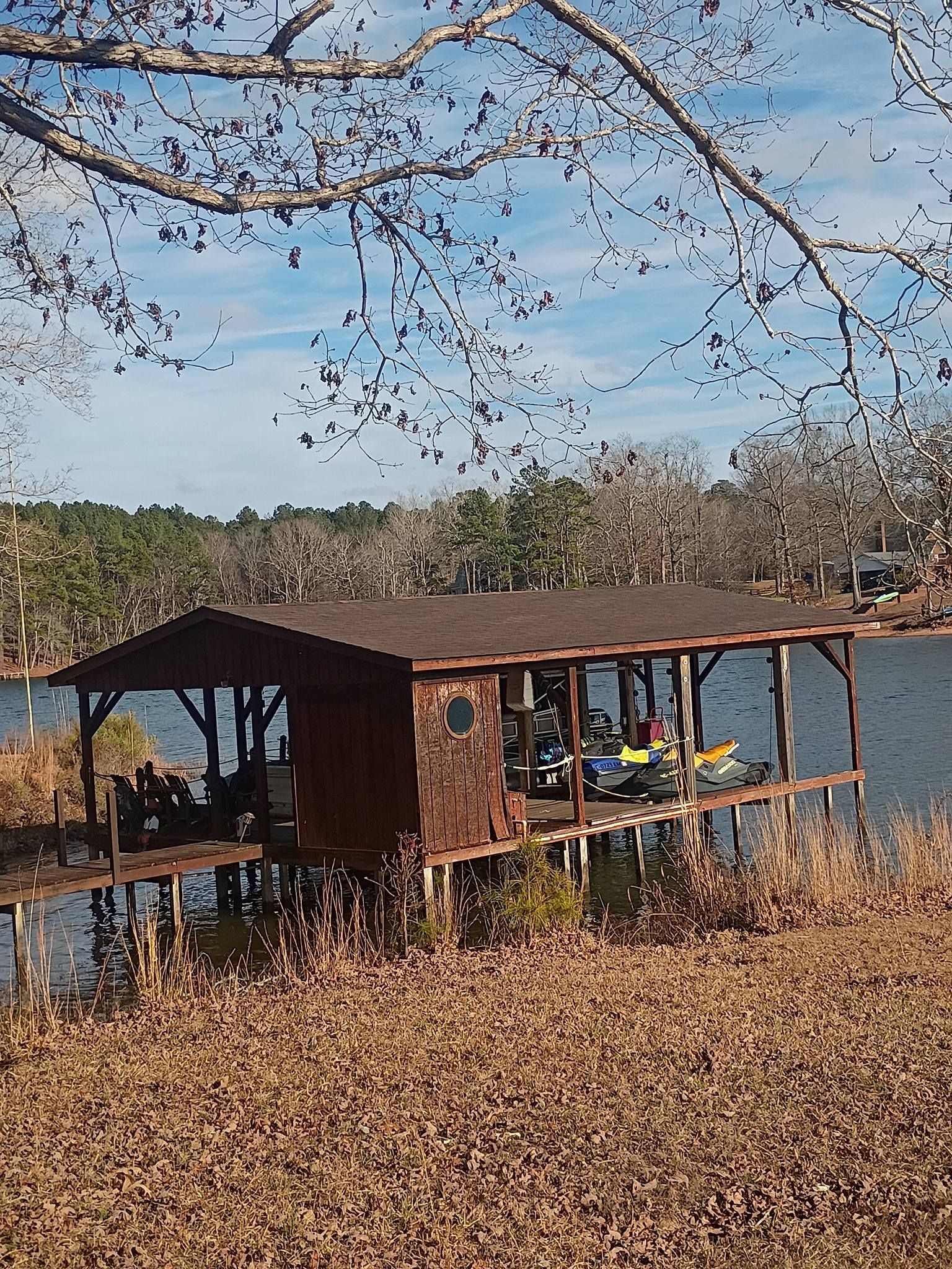 Rustic lakeside cabin on a leaf-covered shore under bare tree branches, with boats visible inside.