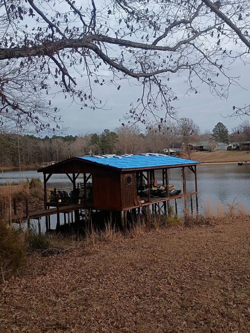 Small lakeside hut with a blue tarp roof under bare tree branches on a cloudy day