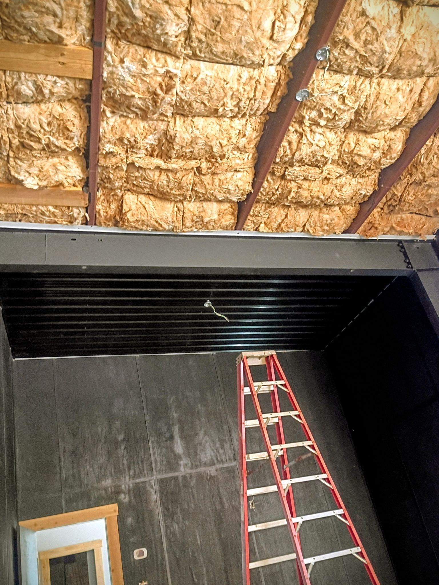 Ladder inside a room with exposed insulation in the ceiling and a black wall panel above a doorway