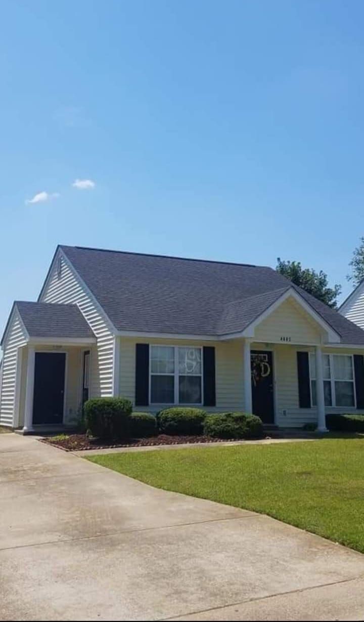 Single-story white house with dark roof, black shutters, front porch, and a driveway under a clear blue sky