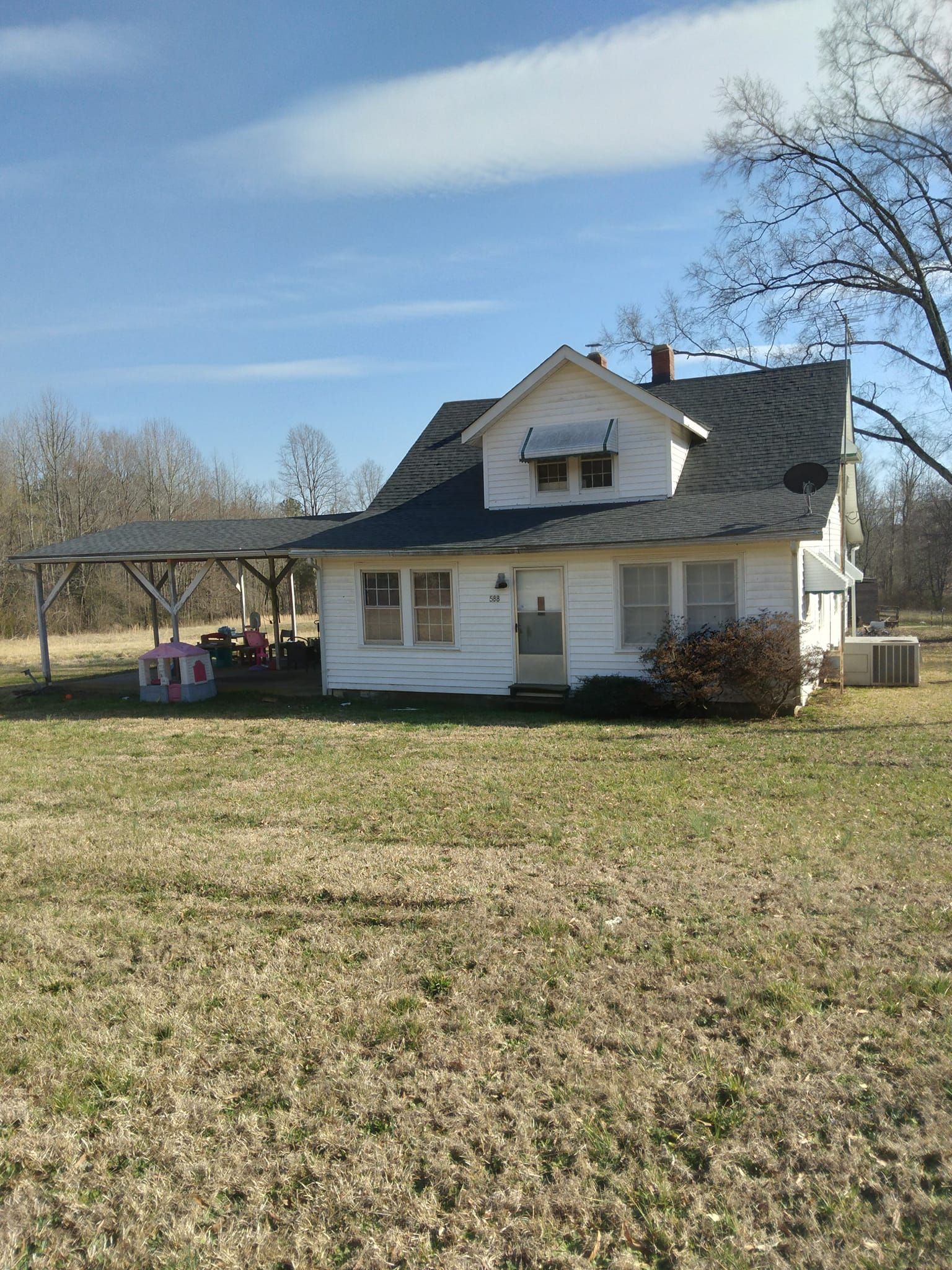 White farmhouse with porch and dormer on a grassy yard under a blue sky, with bare trees nearby.